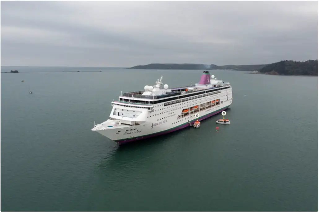 A large white cruise ship, SuperStar Aquarius, from a renowned cruise line is anchored in a calm sea. Surrounded by smaller boats and hills under an overcast sky, it operates with an innovative AI system to ensure smooth sailing and passenger safety.