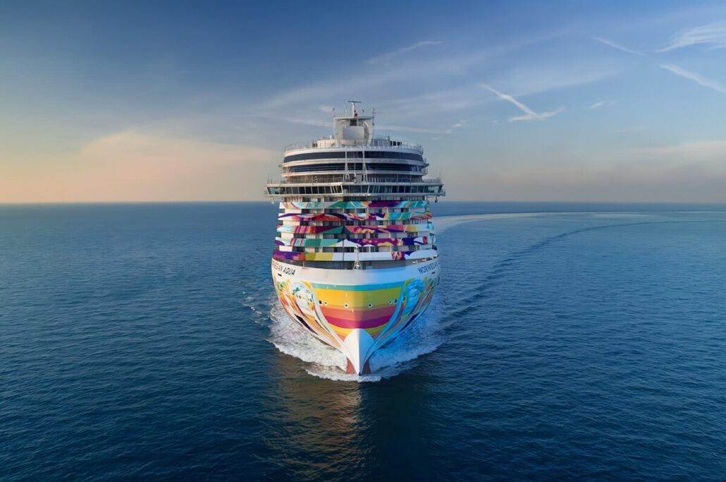 A colorful NCL cruise ship sails forward on a calm ocean under a clear sky. The Prima Plus Cruise Ship's vibrant design features abstract patterns and palm tree motifs. White waves trail behind as it moves through the water, embodying the essence of Norwegian Aqua adventures.