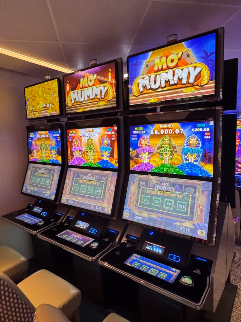 A row of digital slot machines titled "Mo Mummy" stands in a casino setting, featuring a sleek Norwegian Aqua design. The machines display vibrant graphics of pyramids and treasure, while upholstered chairs invite players to enjoy this immersive experience.
