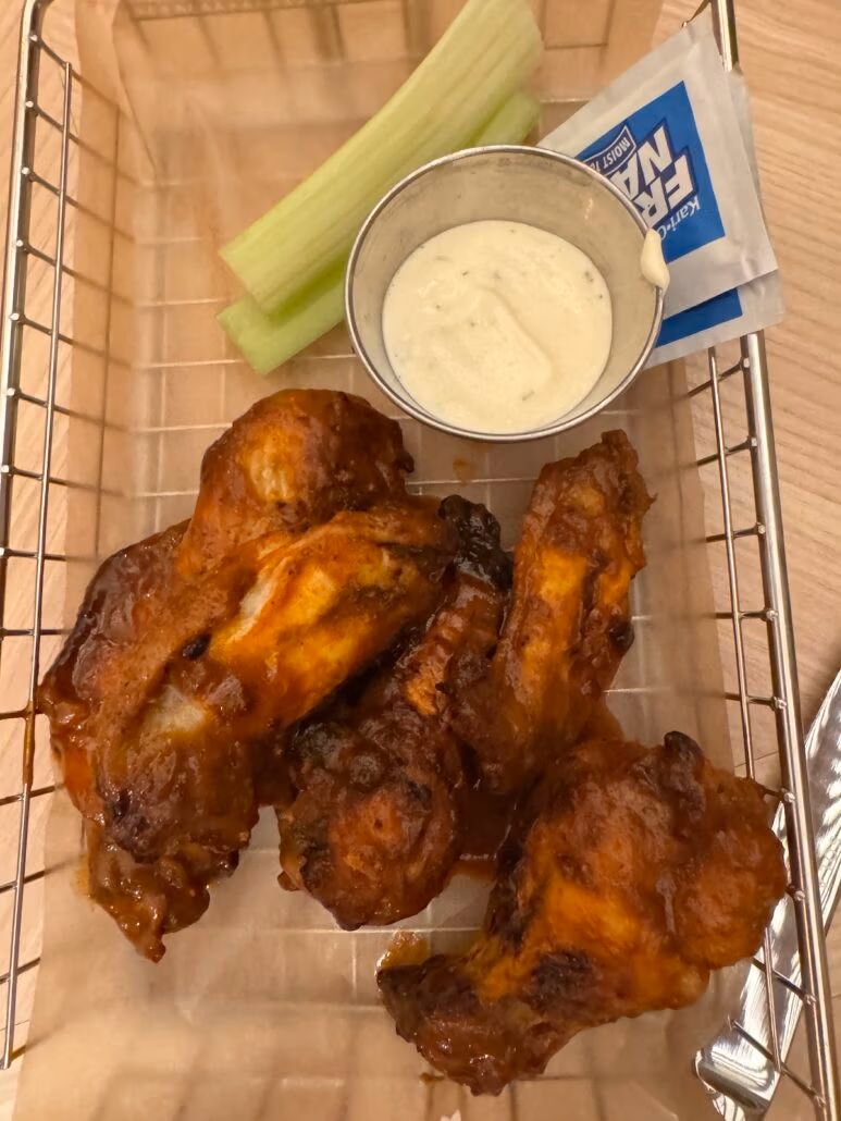 A basket featuring four pieces of buffalo chicken wings, complemented by a sleek metal cup of creamy dipping sauce and a stick of celery. The setup, reminiscent of modern design aesthetics, hints at Norwegian Aqua influences with a butter wrapper partially visible behind the sauce cup.