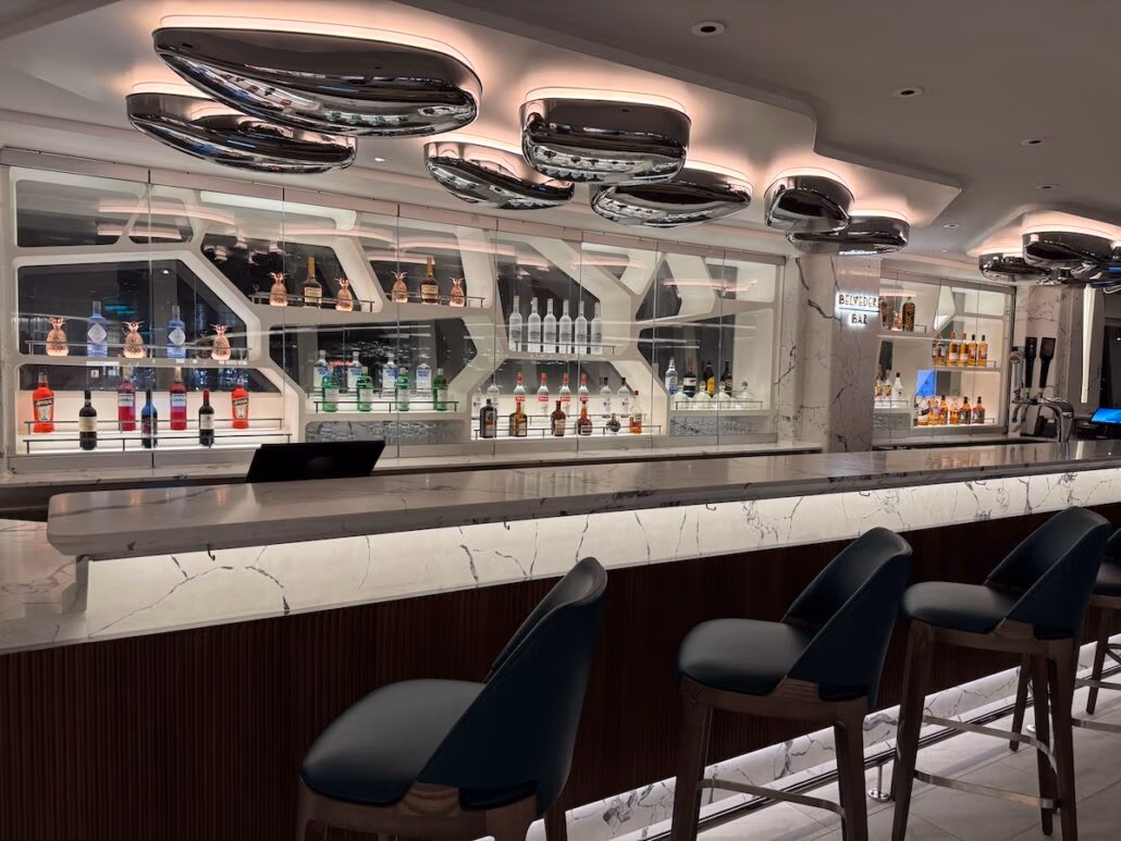 A modern bar with a marble countertop is lined with three teal barstools in the elegant shade of Norwegian Aqua. Behind the counter, shelves display various bottles of alcohol. The ceiling showcases futuristic, metallic light fixtures, enhancing the overall sleek design.