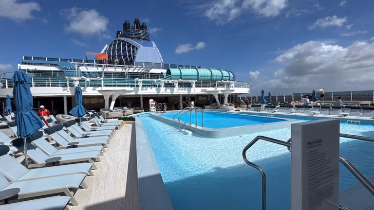 The Norwegian Aqua cruise ship deck boasts a stunning design, featuring a large swimming pool surrounded by lounge chairs and blue umbrellas. Under the clear sky, a vigilant lifeguard keeps watch, while in the background, the ship's elegant structure stands proudly.
