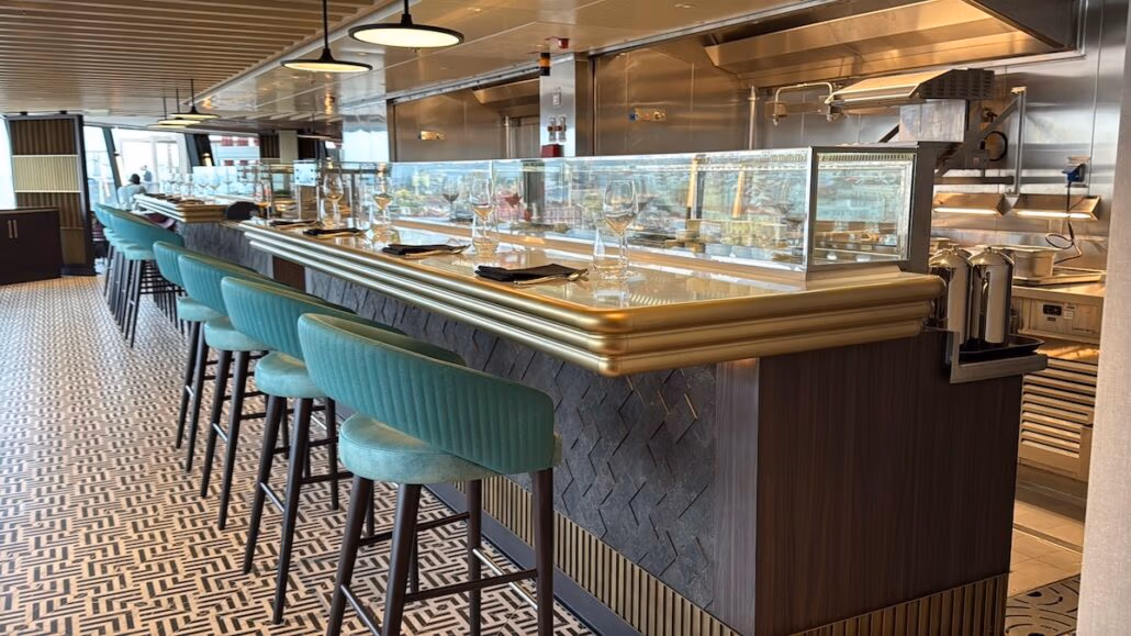 A modern bar-style dining area boasts high Norwegian Aqua chairs lined along a counter. The elegant gold-trimmed counter with a glass display exudes sophistication. The surrounding decor includes patterned flooring and hanging lights, while kitchen equipment is visible in the background.