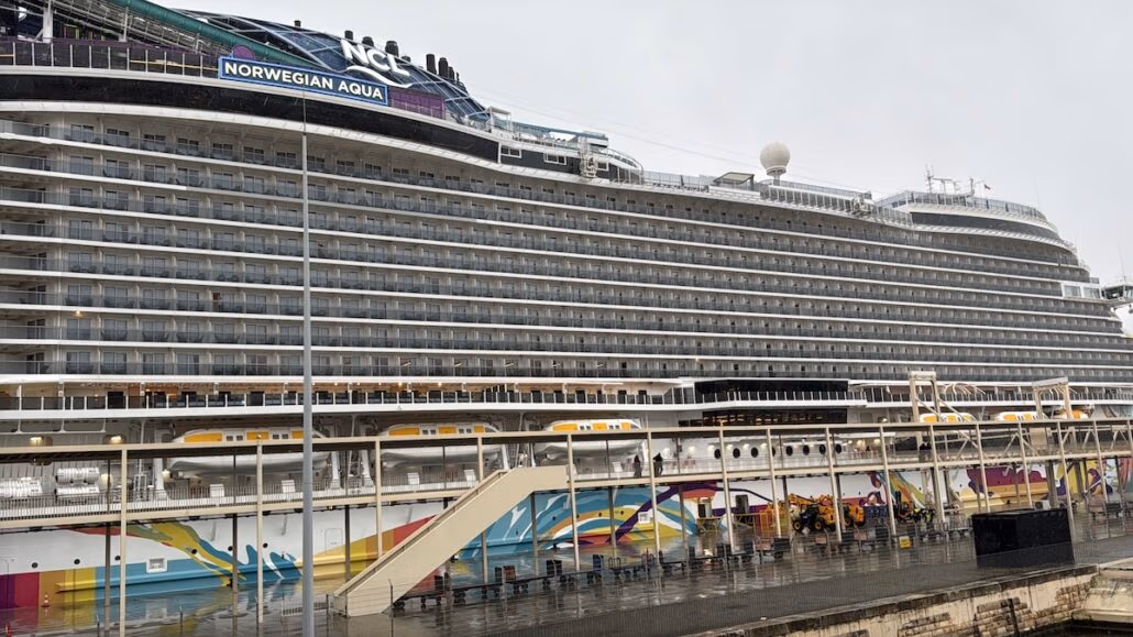 The large cruise ship, Norwegian Aqua, docked at the port under a cloudy sky, showcases its exquisite design. The vessel displays multiple decks with rows of balconies, while various colorful patterns adorn the lower hull.