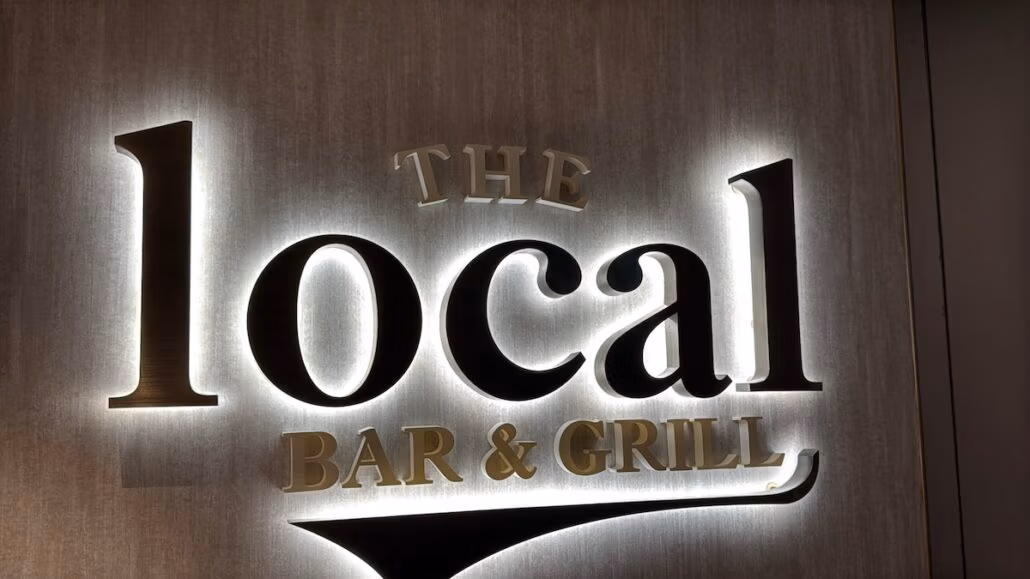 A sign reading "The Local Bar & Grill" with illuminated lettering stands out against the textured wall, reflecting a touch of Norwegian Aqua in its sleek design. Perfectly capturing the essence of dining, it invites patrons for an unforgettable experience.