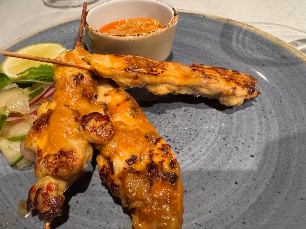 Grilled chicken skewers rest elegantly on a Norwegian Aqua plate, paired with a fresh salad garnish and a small bowl of dipping sauce. A lime wedge adds a zesty design touch, perfectly enhancing this delightful dining presentation.