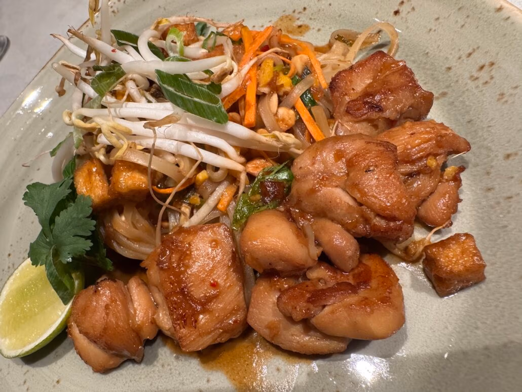 A plate of stir-fried chicken pieces with bean sprouts, green onions, and sliced vegetables rests elegantly on a round, light green dish in true Dining design style. A piece of lime and a sprig of cilantro add freshness, completing this visually striking culinary presentation.