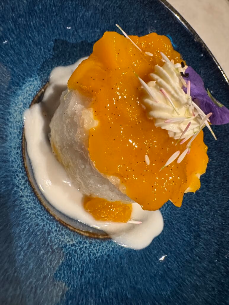 A dessert masterpiece featuring rice topped with vibrant orange mango chunks and a dollop of cream, garnished with a purple petal and white sprinkles. Served on a Norwegian Aqua blue plate, the presentation is elevated by a swirl of white sauce underneath, perfect for sophisticated dining.