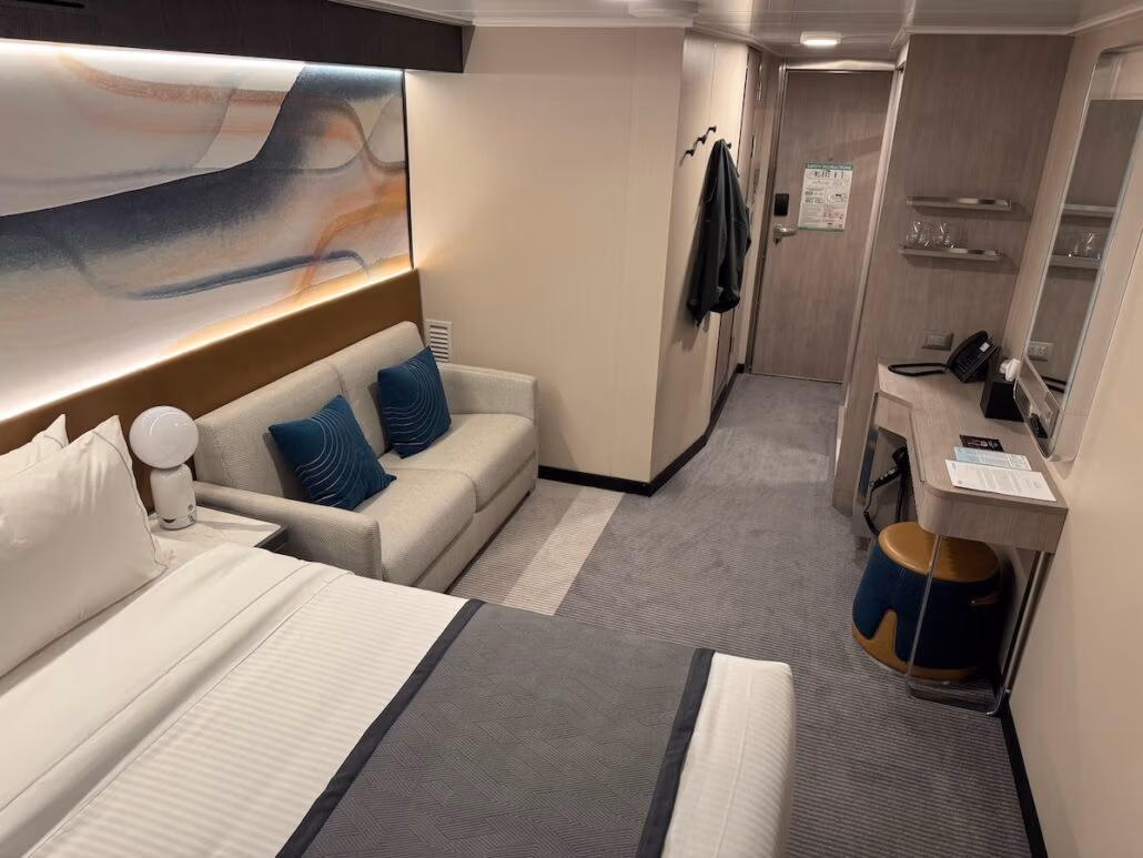 A neatly arranged hotel room features a bed with white linens, a gray sofa adorned with blue cushions reminiscent of Norwegian Aqua, and a small desk with a stool. A jacket hangs on the wall near the entrance, while light-colored walls display abstract artwork above the sofa.