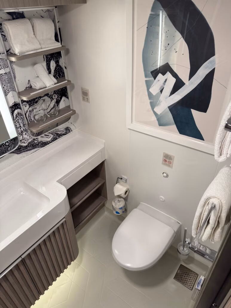 Modern bathroom with a wall-mounted toilet, a white basin on a vanity with wooden drawers, and shelves holding towels. Abstract art hangs on the wall, reflecting in the mirrored backsplash, while gray herringbone floor tiles evoke the calm of Norwegian Aqua.