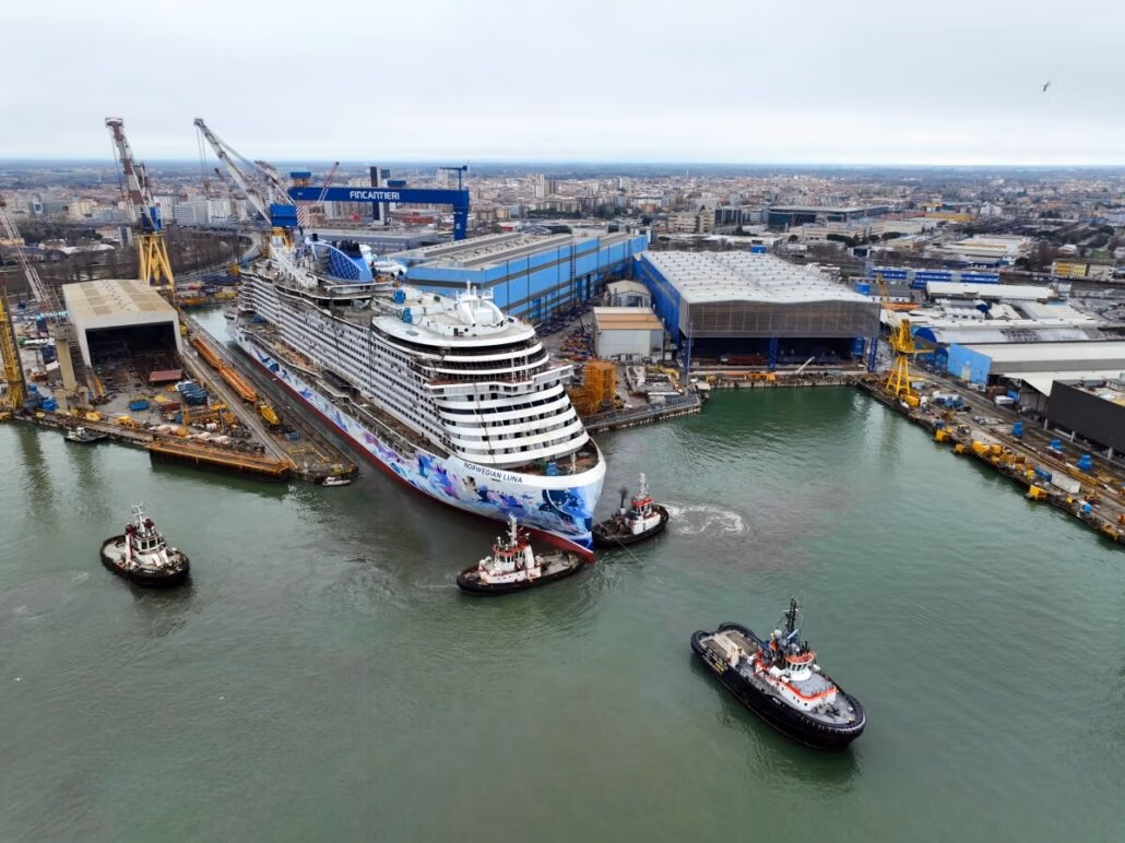 Aerial view captures the 2026 launch of the Norwegian Luna, surrounded by four tugboats near a bustling shipyard. The cruise ship floats out under a cloudy sky with industrial buildings and cranes nearby, as the cityscape forms a distant backdrop.