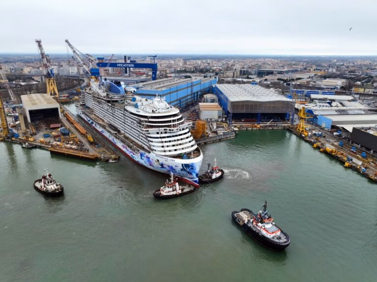 Aerial view captures the 2026 launch of the Norwegian Luna, surrounded by four tugboats near a bustling shipyard. The cruise ship floats out under a cloudy sky with industrial buildings and cranes nearby, as the cityscape forms a distant backdrop.