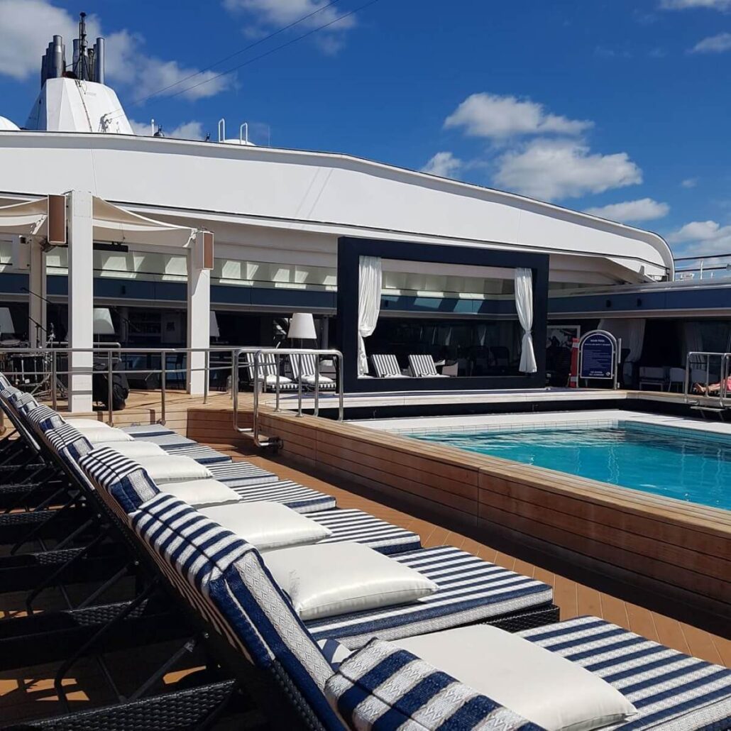 A serene pool deck on a Carnival Corp cruise, featuring lounge chairs with striped cushions lined up in rows. A beautiful swimming pool is surrounded by a wooden deck, while white curtains elegantly frame sections of the ship's structure under a clear sky with a few clouds.