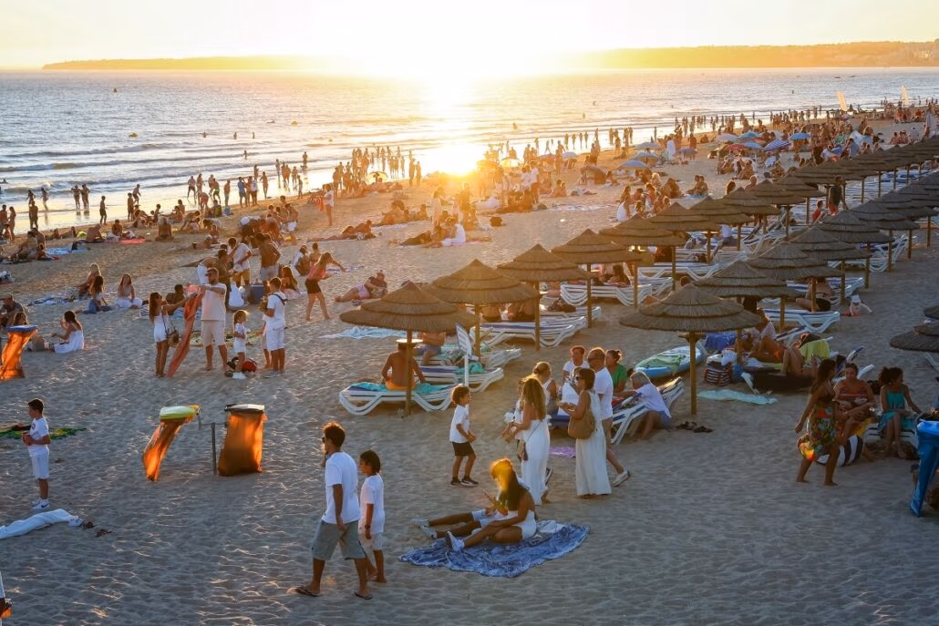 At sunset, on a sandy beach in Portugal, people lounge on sunbeds under straw umbrellas while others stroll or sit on the sand. The ocean stretches under a clear sky as the sun kisses the horizon, despite the recent ban on bikinis looming over this serene setting.