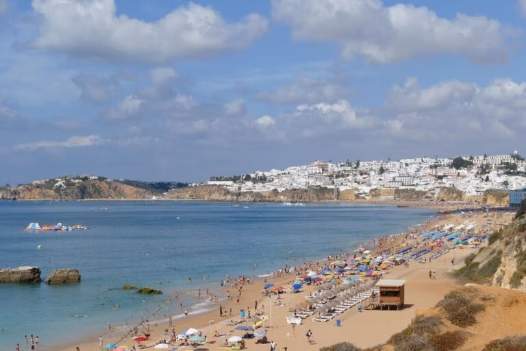 A scenic view of a sandy beach in Portugal, lined with colorful umbrellas and numerous beachgoers in bikinis. The turquoise sea is calm, with a few boats in the distance. In the background, a town with white buildings sits atop a hill under a partly cloudy sky.