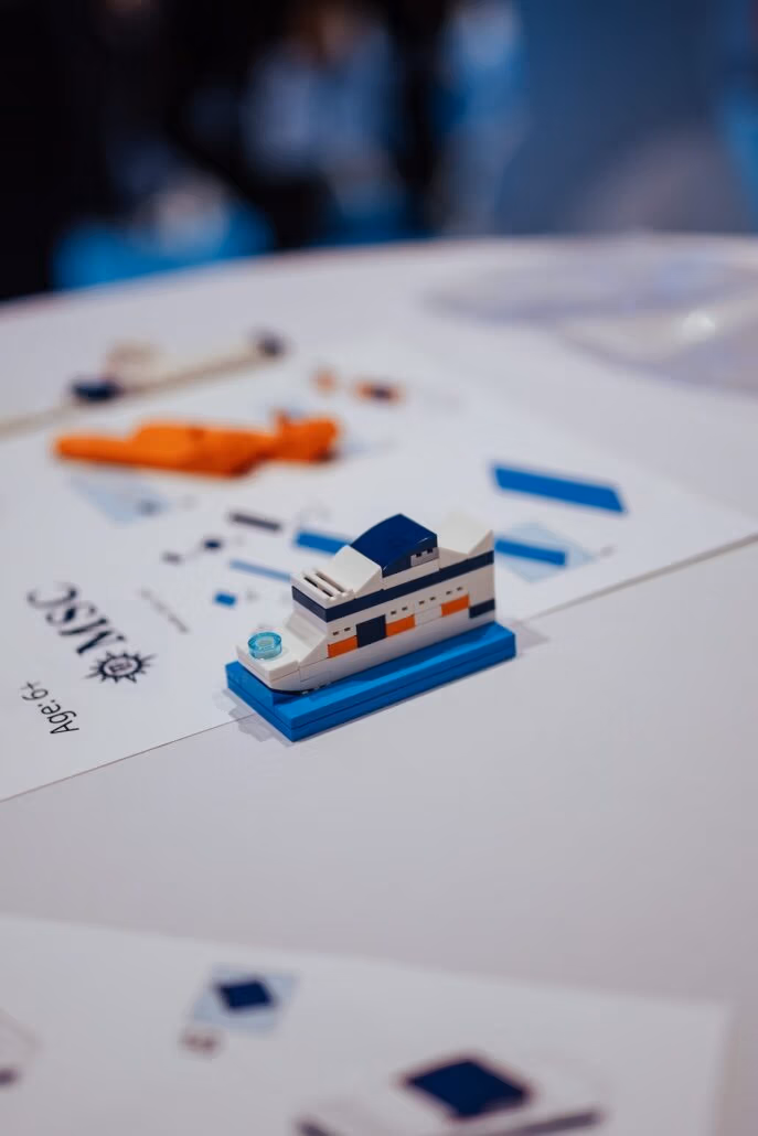 A small block model of an MSC ship sits on a table, surrounded by papers and instructions. The LEGO display features white, blue, and orange pieces. A pen and other materials are visible in the background, adding to the creative shipbuilding atmosphere.
