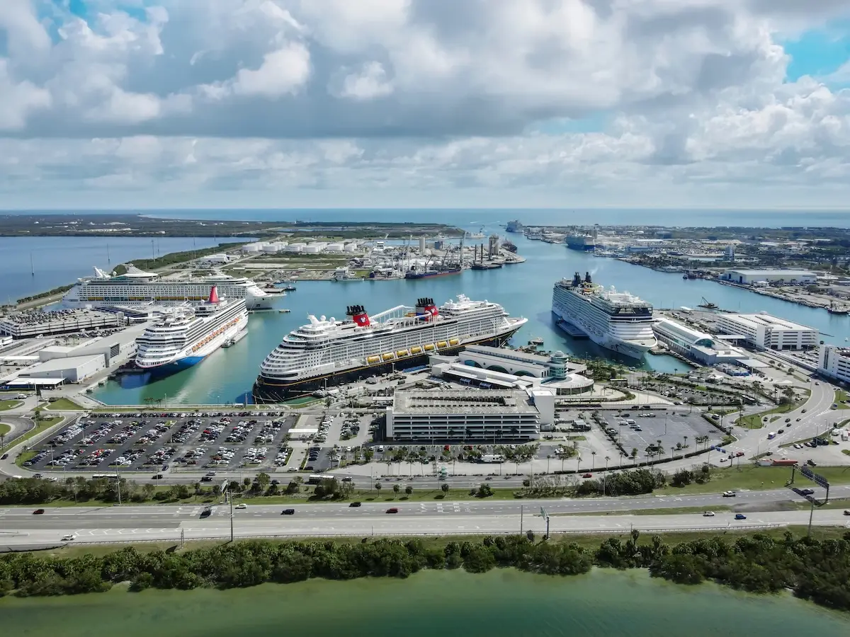 Aerial view of Port Canaveral, with several large cruise ships docked at terminals, parking lots filled with vehicles, and various port buildings bustling with cruisers under a partly cloudy sky.