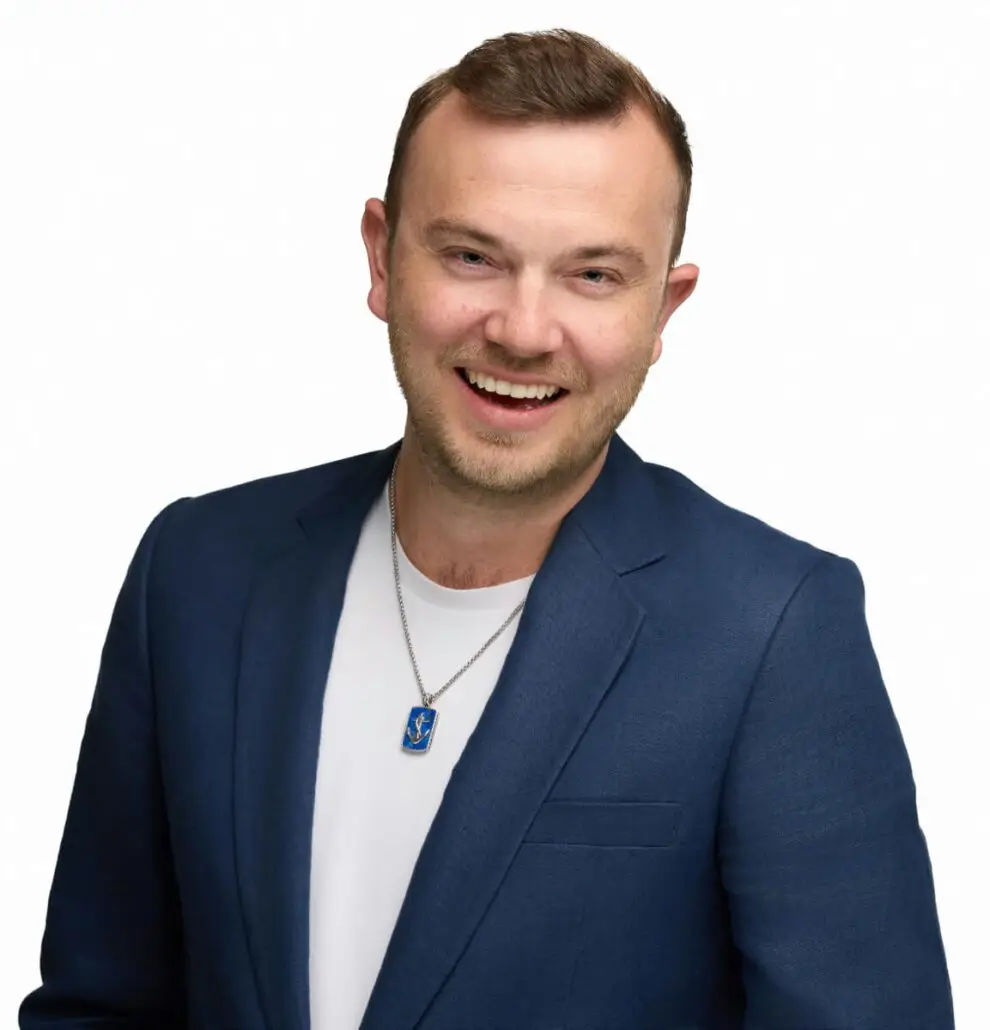 A man with short light brown hair and a trimmed beard wears a blue blazer over a white shirt and a blue pendant necklace. He is smiling at the camera, ready to announce the 2025 Carnival Cruise Director schedule against a plain white background.