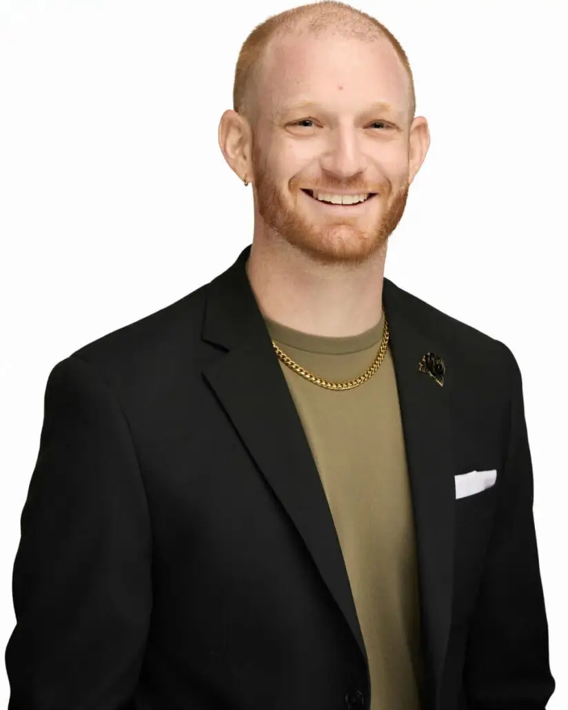 A person with short red hair and a beard is smiling while wearing a black blazer over an olive green shirt, as if ready for the 2025 Carnival Cruise Director Schedule, standing confidently against a plain white background.