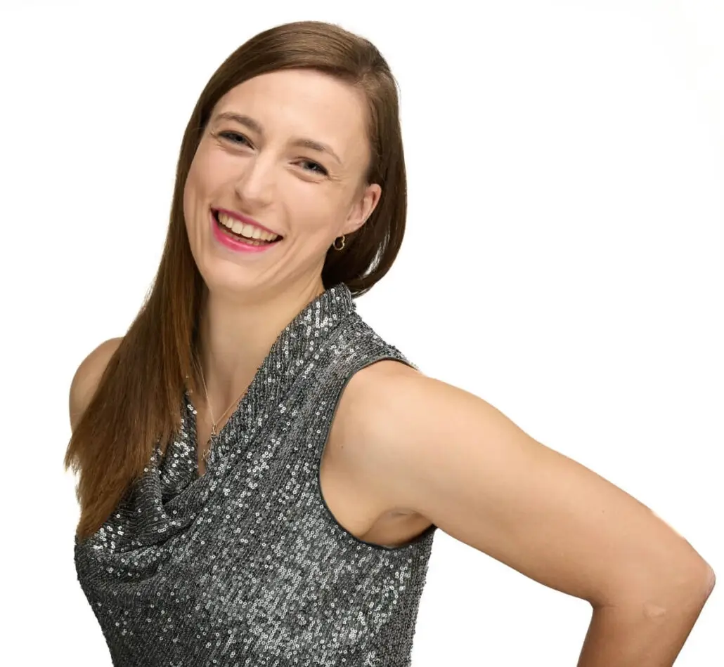 A woman with straight brown hair, smiling in a sleeveless silver sequined top, stands confidently against a white background—ready for her role as a cruise director on the Carnival Cruise 2025 schedule.