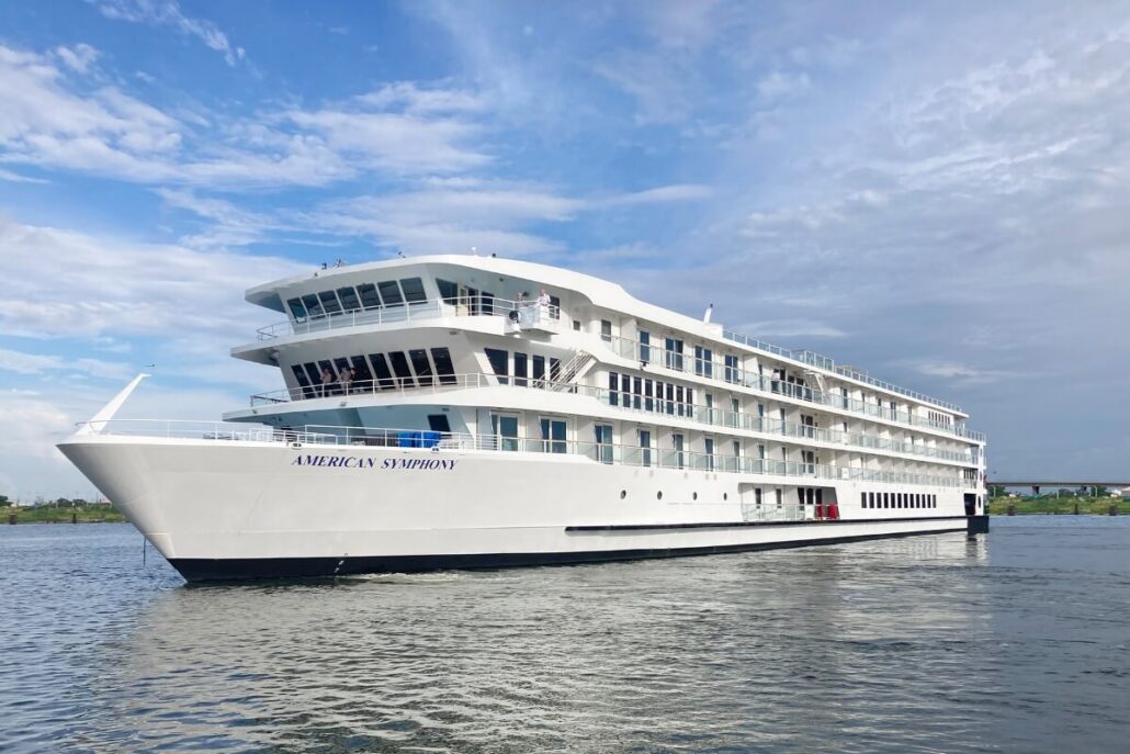 A large white river cruise ship named "American Symphony," part of leading Mobile cruise companies, sails on calm water under a partly cloudy sky, with multiple decks visible and passengers relaxing on the upper level.