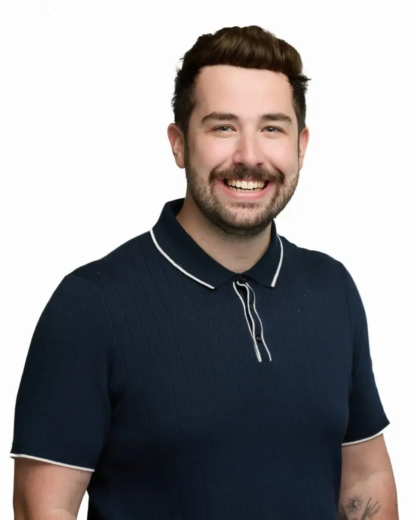 A person with short brown hair, a mustache, and beard smiles at the camera. They wear a dark navy short-sleeve polo with white trim. The plain white background gives a polished look—perfect for the 2025 Carnival Cruise director schedule announcement.
