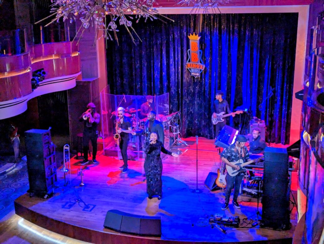 A band performs on stage aboard Holland America’s Koningsdam, with a singer up front, horn players to the left, drummer and bassist in back, and two guitarists to the right. The BB King’s Blues Club logo shines against dark curtains—true Alaska cruise vibes.