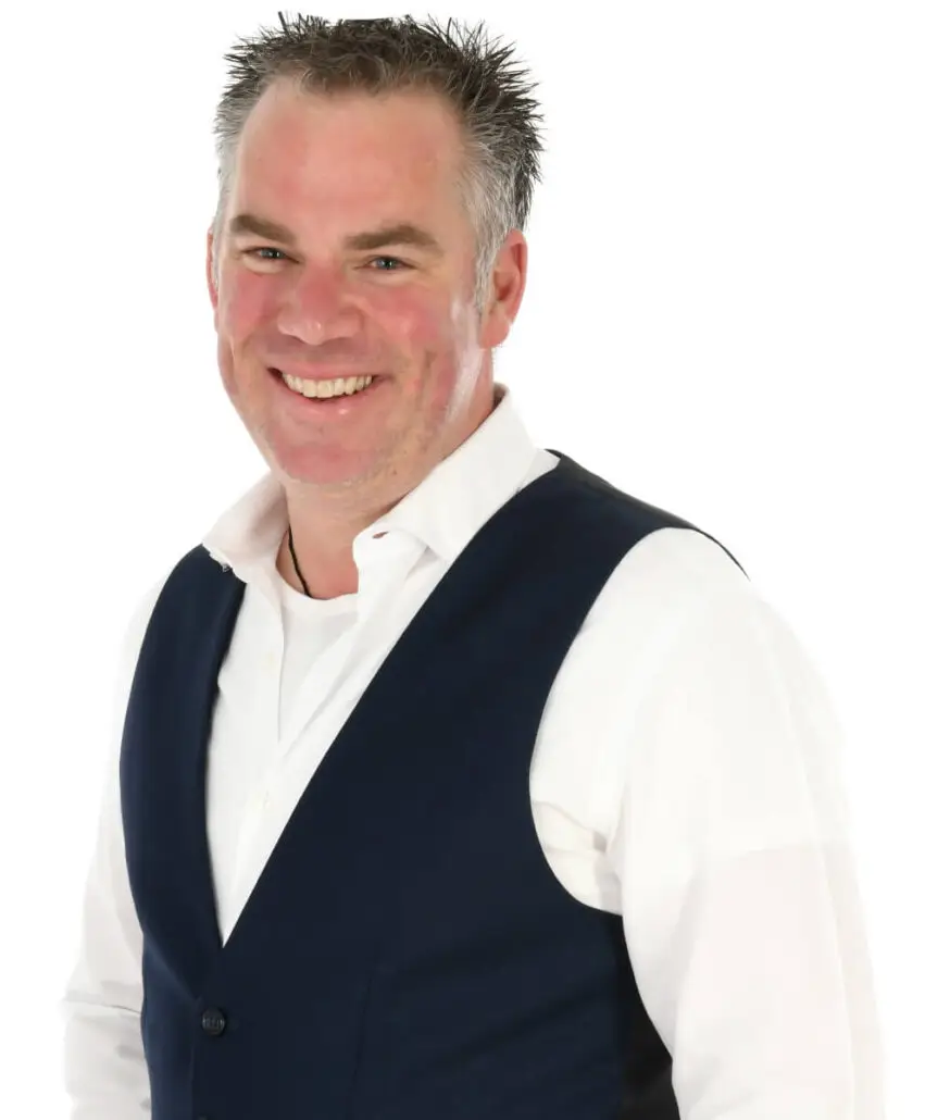 A man with short, spiked gray hair is smiling at the camera. He is wearing a white dress shirt and a dark blue vest, looking every bit the professional cruise director you'd expect from the Carnival Cruise Director Schedule for 2025. The background is plain white.