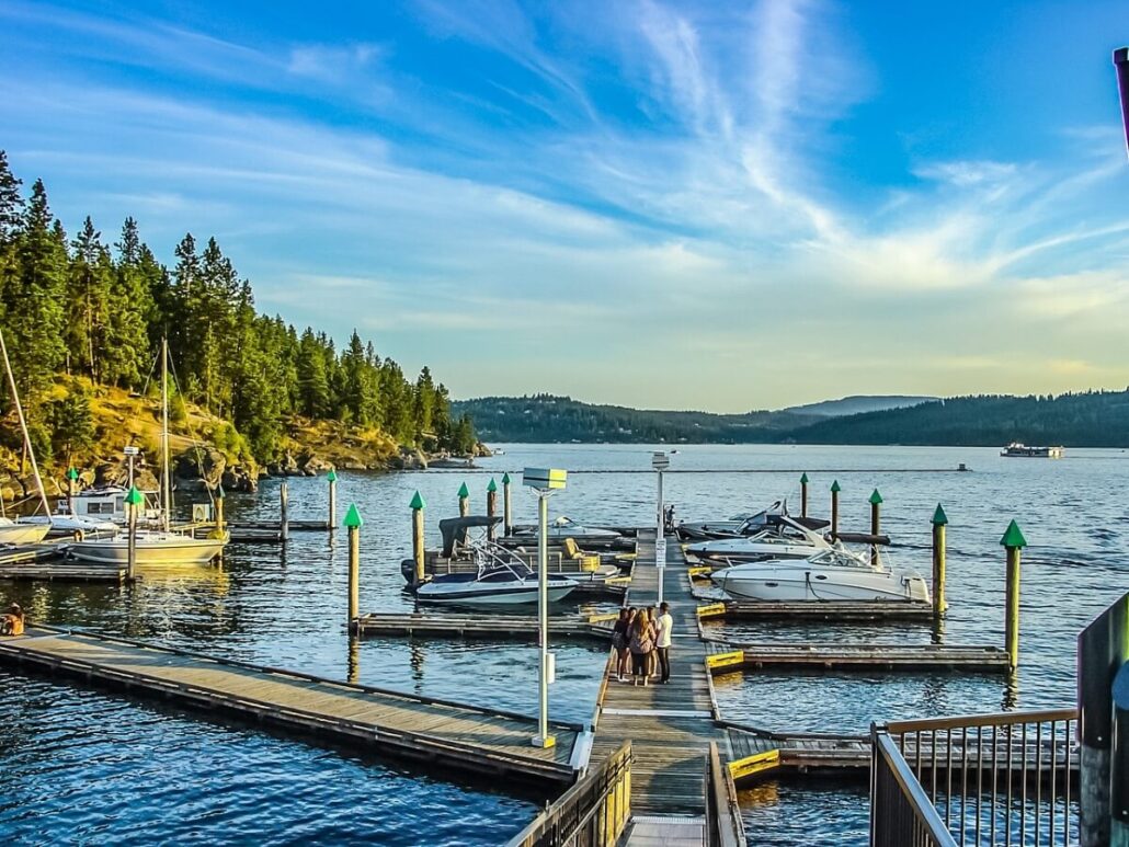 A wooden dock with several motorboats and a sailboat moored on Lake Travis, surrounded by forested hills under a blue sky. Two people sit together at the end of the dock, enjoying the peaceful views near Lake Travis boat slips.