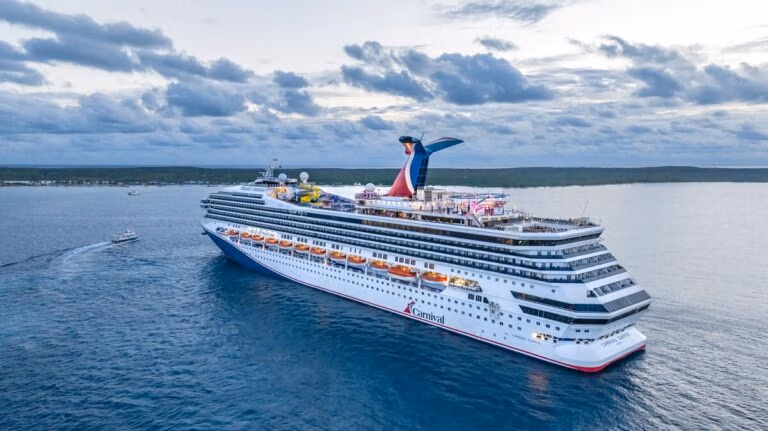 A large Carnival cruise ship sails on calm blue water near a distant coastline under a partly cloudy sky. The ship, resembling a Michigan cruise ship, has multiple decks, lifeboats, and a distinctive red and blue funnel.