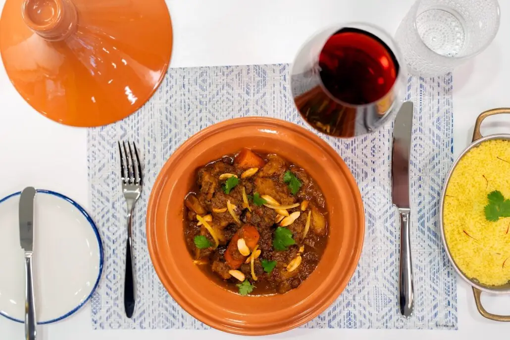 Overhead view of a table setting at a new brunch venue with a Moroccan tagine in a clay pot, red wine, covered dish, plate with utensils, and casserole of yellow couscous garnished with cilantro—perfect for Celebrity Xcel's all-day entertainment vibes.