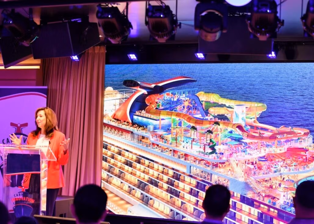 A person stands at a podium giving a presentation. Behind them, a large screen showcases a Carnival cruise ship adorned with vibrant water slides and recreational areas on deck, enticing the audience to consider the benefits of joining the VIFP Program for an unforgettable experience at sea.