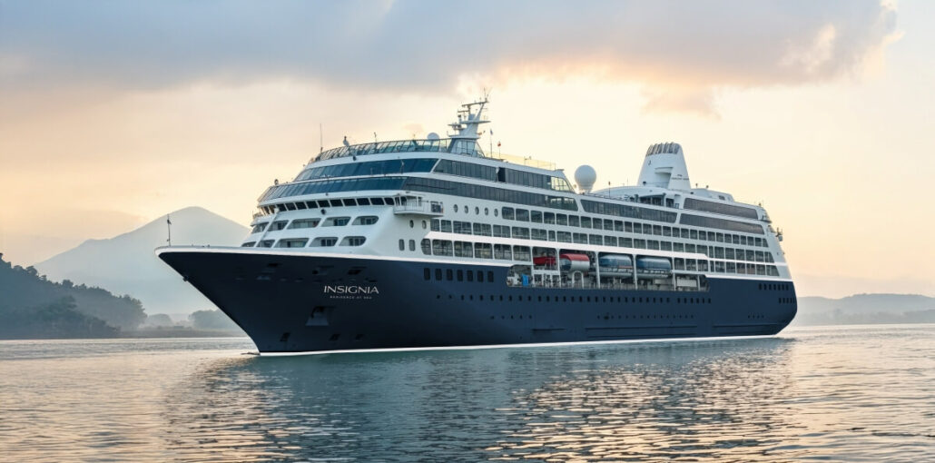 A large cruise ship from a renowned cruise line, with a dark blue hull and white upper decks, sails on calm waters. The overcast sky allows a hint of sunlight to break through, while mountains faintly loom in the background.