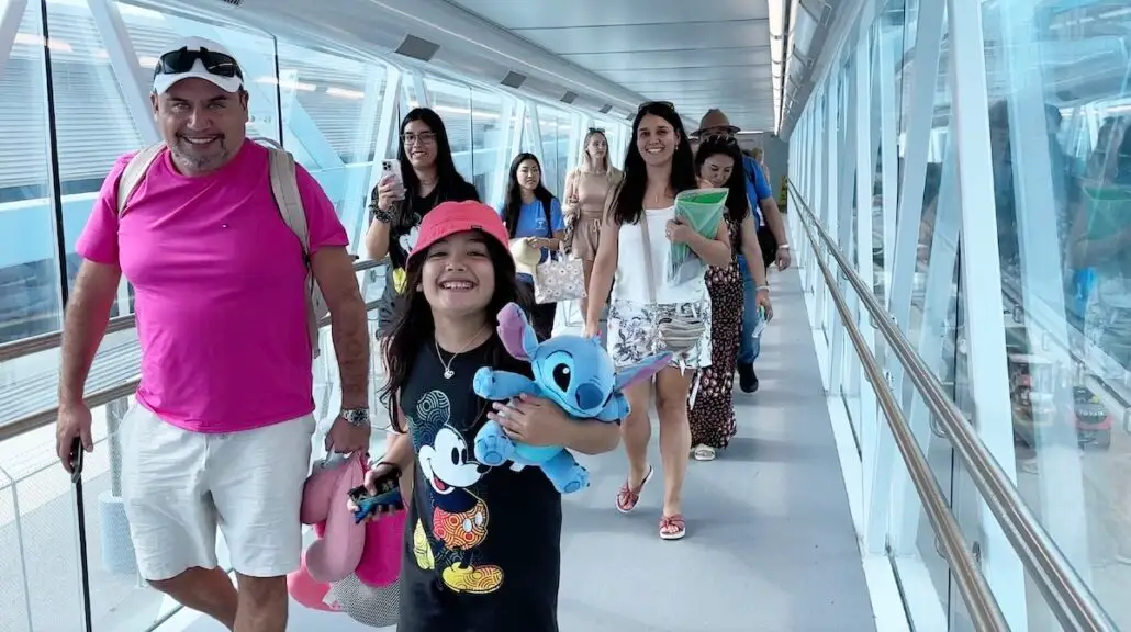 A group of cruisers, including a smiling child holding stuffed toys and adults carrying bags, walk down a glass-enclosed hallway, possibly at Port Canaveral or a cruise terminal.
