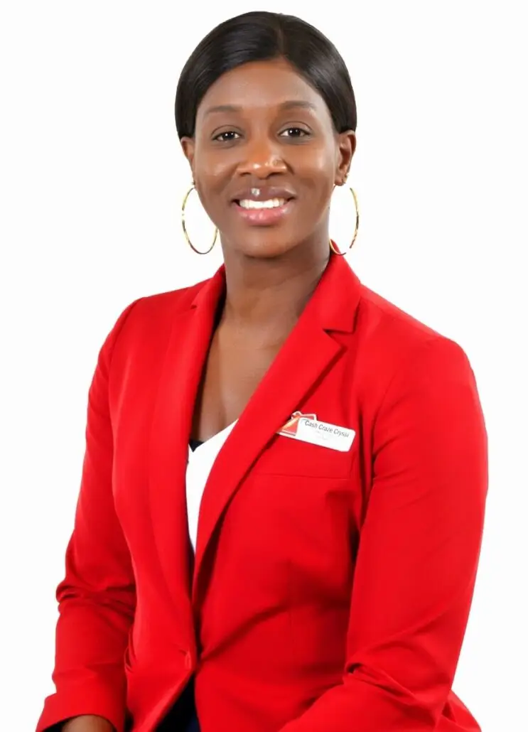 A woman wearing a red blazer and large hoop earrings sits facing the camera, smiling. With straight, dark hair pulled back and a name badge on her blazer, she appears ready for her 2025 role as a Carnival cruise director. The background is plain white.
