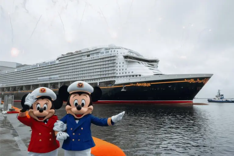 Two people in Mickey and Minnie Mouse costumes dressed as ship captains stand on a dock before the Mega Ship Disney Adventure, a proud addition to the Disney Cruise Line, with overcast skies and faint fireworks in the background.