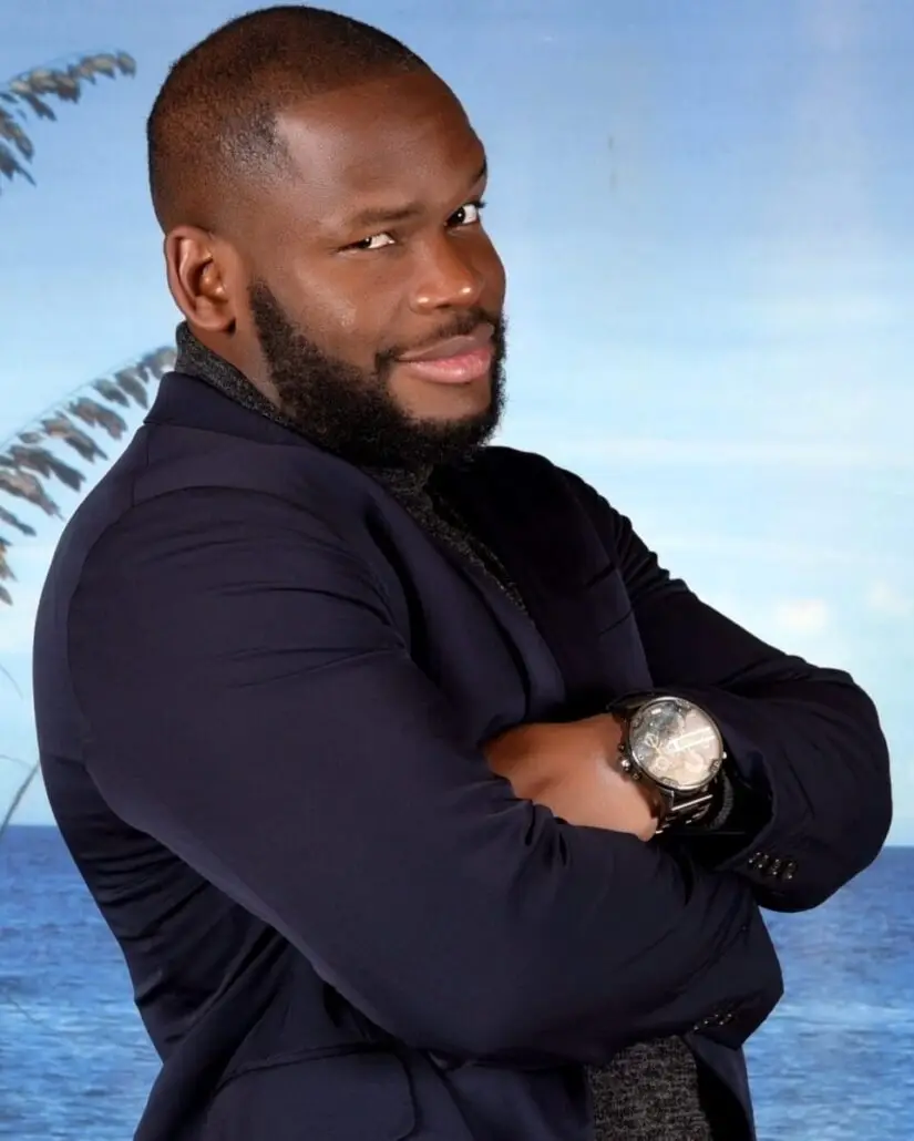 A man with a short beard and closely shaved hair stands with arms crossed, wearing a dark blazer and watch, in front of a blue backdrop evoking the sea and sky—perfect for a 2025 Carnival Cruise Director schedule reveal.