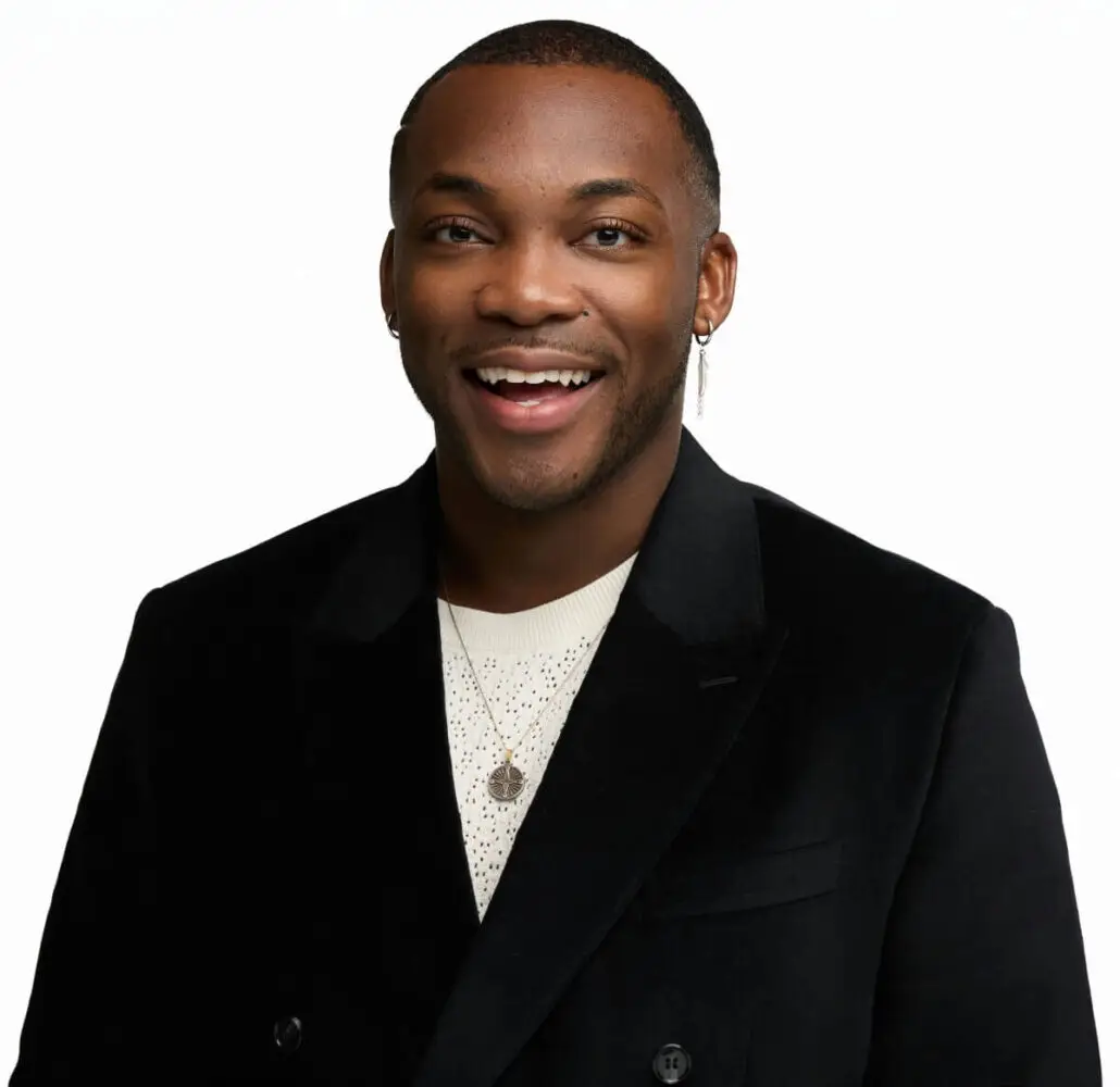 A person wearing a black blazer over a white textured shirt, smiling with their mouth slightly open. They have short hair and are wearing small hoop earrings and layered necklaces—ready to announce the Carnival Cruise Director Schedule. The background is plain white.