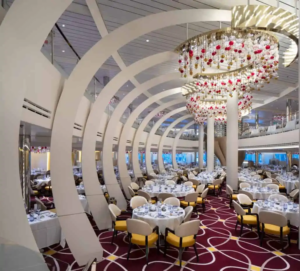 A spacious, modern dining room aboard Holland America’s Koningsdam features round tables set for meals, cream-colored chairs, large arching beams, a colorful patterned carpet, and chandeliers decorated with red and clear glass ornaments.
