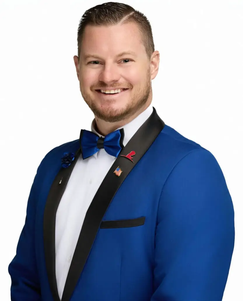 A man with short light brown hair and a beard, dressed as a cruise director in a bright blue suit jacket with black lapels, a blue bow tie, and a white shirt, smiles at the camera—perfect for the 2025 Carnival Cruise Director Schedule.