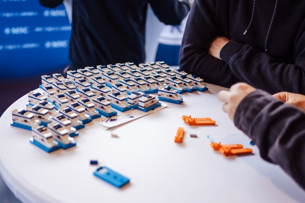 Two people are assembling small boat models on a round table, creating an impressive LEGO display. Numerous completed miniature ships are arranged in rows. Orange and white blocks are scattered on the table. The scene is indoors with a blurred background, capturing their creative focus.