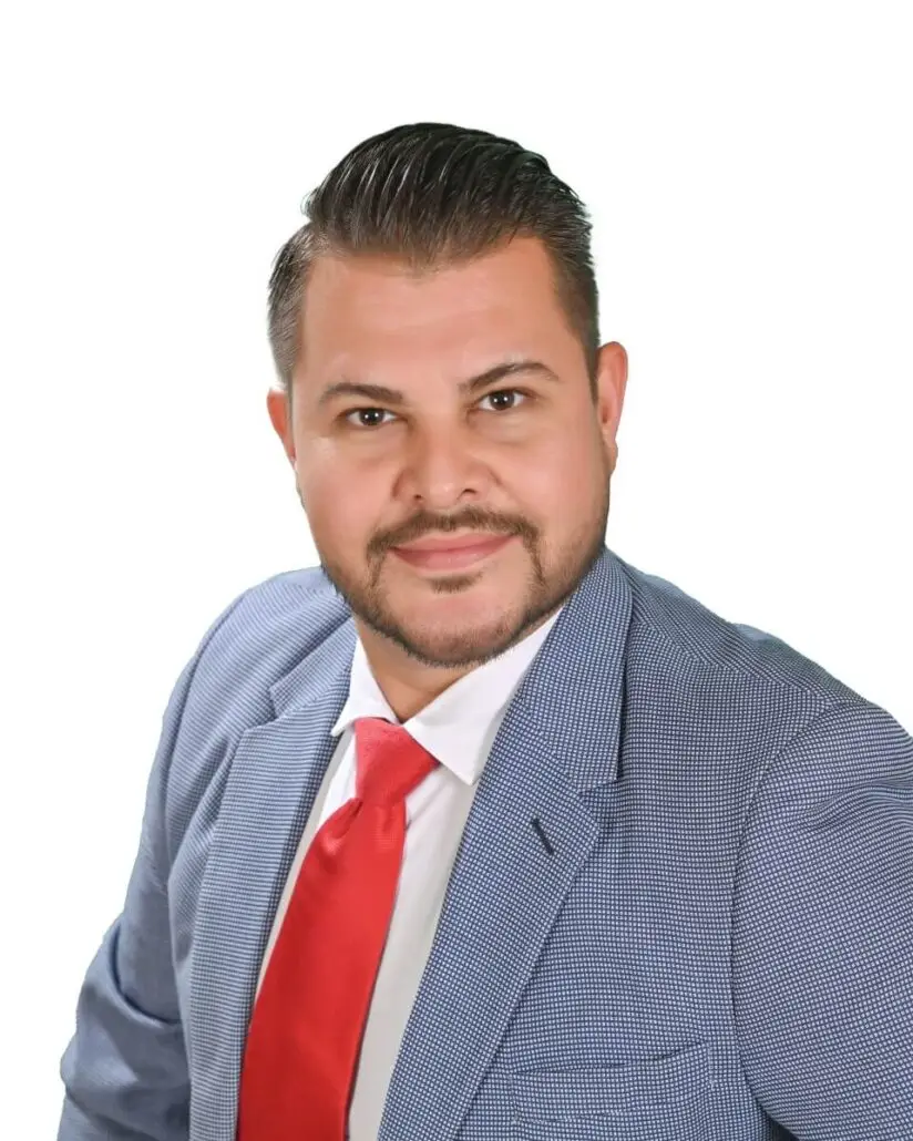 A man with short dark hair and a trimmed beard—dressed in a light blue textured suit jacket, white shirt, and bright red tie—poses against a plain white background, reminiscent of a Carnival Cruise Director Schedule 2025 headshot.