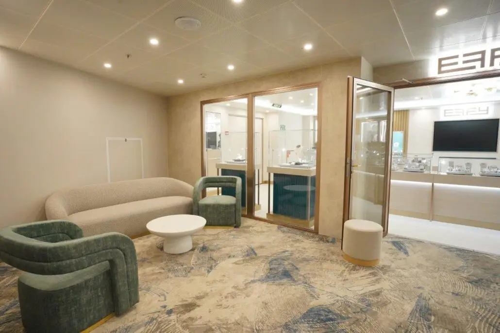 A modern waiting area with a beige sofa, two green armchairs, and a round white coffee table evokes the relaxing ambiance of a spa. Glass doors lead to a brightly lit store with display cases and a large screen.