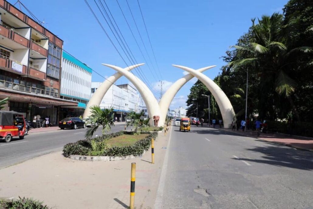 A bustling urban street invites tourism with its large ivory tusk-shaped arches spanning across, nestled between plant-lined sidewalks. Cruise passengers and locals intermingle among cars, with towering multi-story buildings lining the scene under a clear blue sky.