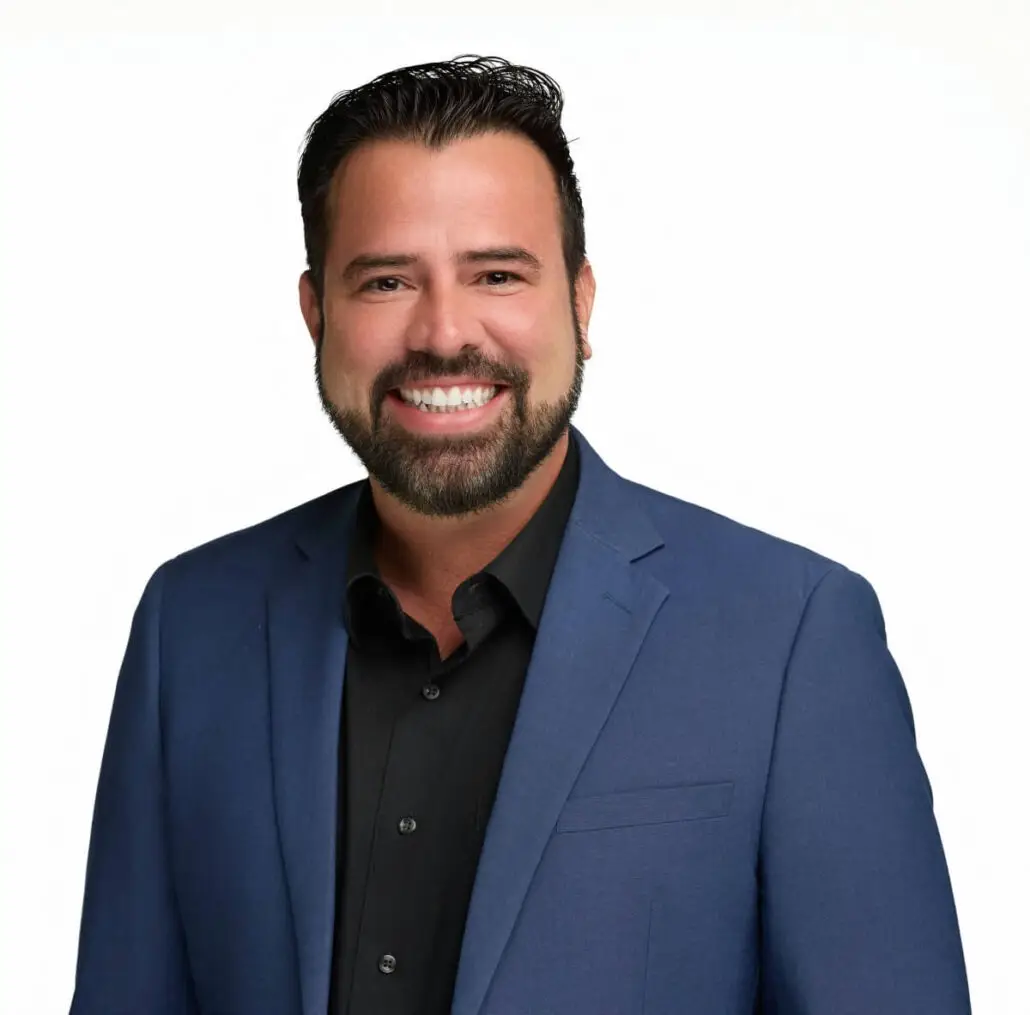 A man with dark hair and a beard is smiling at the camera. Dressed in a dark blue suit jacket over a black shirt, he could easily be mistaken for a cruise director posing for the 2025 Carnival Cruise Director Schedule against a plain white background.