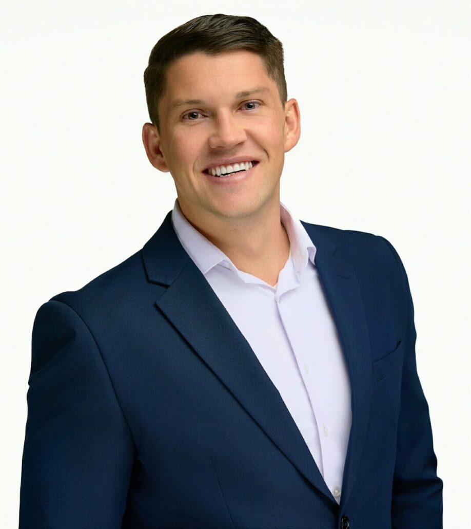 A man with short brown hair is smiling and wearing a dark blue suit jacket over a white dress shirt, standing against a plain white background, ready for his 2025 role as a cruise ship director.