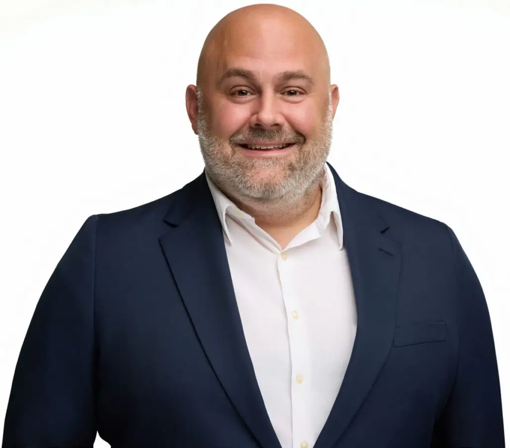 A bald man with a beard, dressed in a navy blue suit jacket and white dress shirt, smiles at the camera against a plain white background—ready to announce the 2025 Carnival Cruise Director Schedule for your favorite ship.