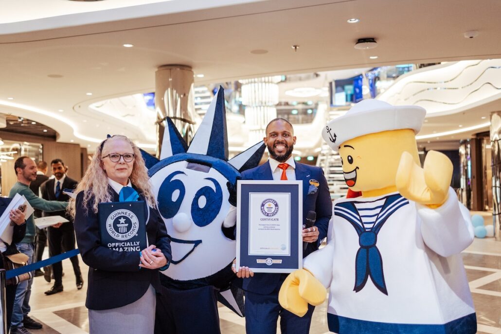A man in a suit proudly holds a Guinness World Records certificate, flanked by a woman with glasses holding an MSC branded plaque and people in mascot costumes like the Statue of Liberty and a sailor, all celebrating near a stunning LEGO display inside a bustling shopping mall.
