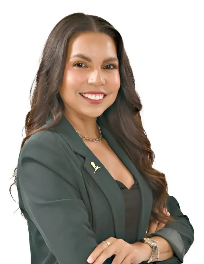 A woman with long brown hair, wearing a dark green blazer and black shirt, smiles with arms crossed. A gold pin on her lapel hints at her role as a Cruise Director for the 2025 Carnival Cruise Director Schedule, against a plain white background.