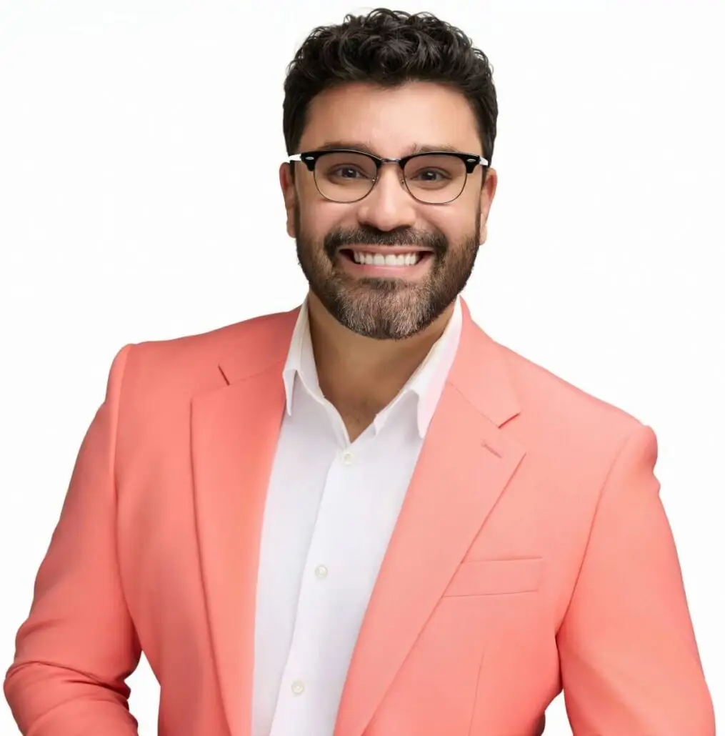 A man with dark hair, a beard, and glasses is smiling. He is wearing a coral pink blazer over a white dress shirt—ready to announce the Carnival Cruise Director Schedule for Carnival Cruise 2025 against a plain white background.